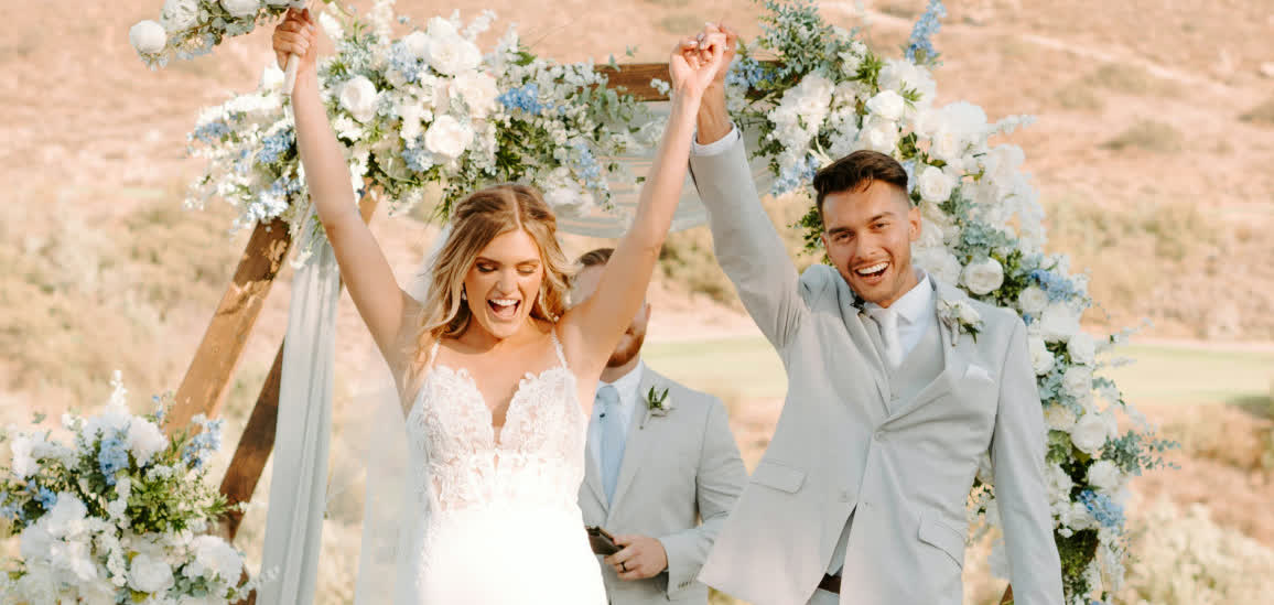 Newlyweds celebrating with raised hands beneath a floral ceremony arch